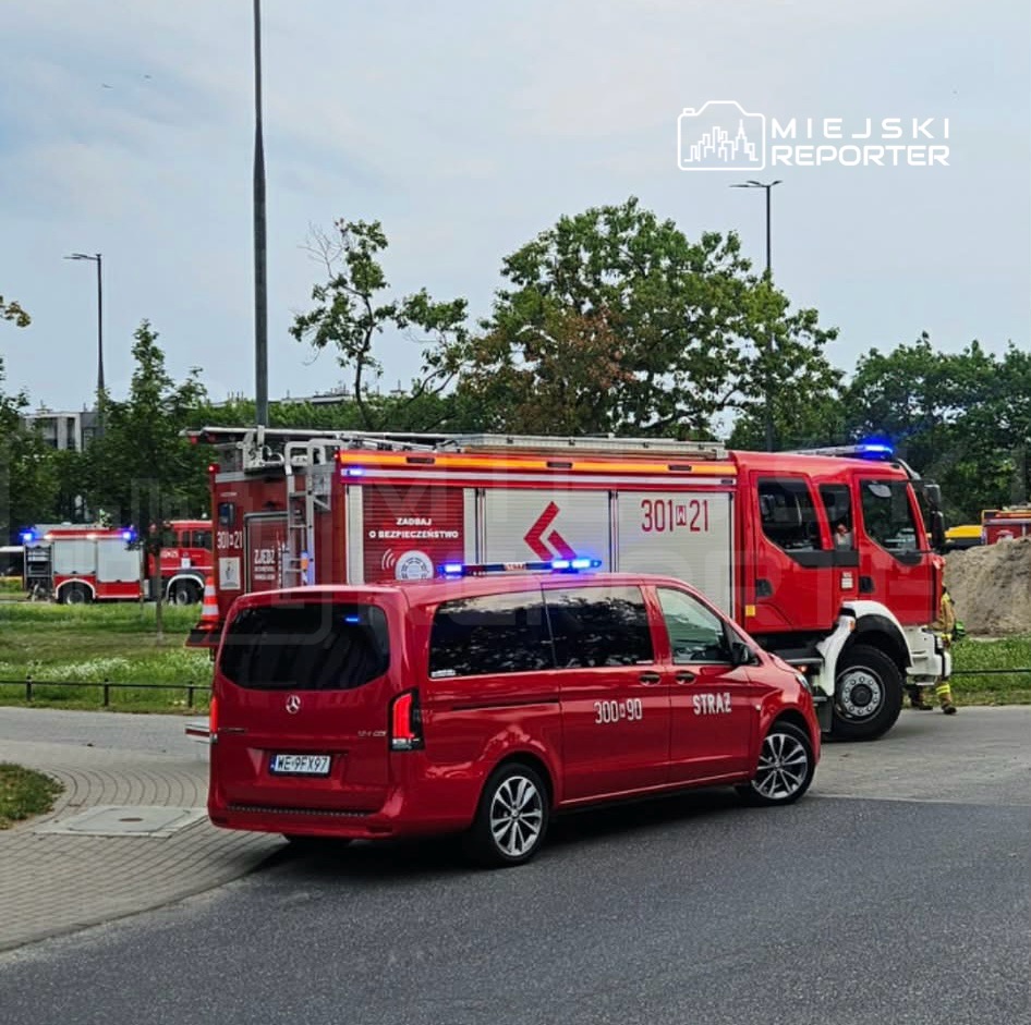 Czerwony wóz strażacki oraz samochód służbowy z numerem 300-90 zatrzymane na skrzyżowaniu w pobliżu zieleni.