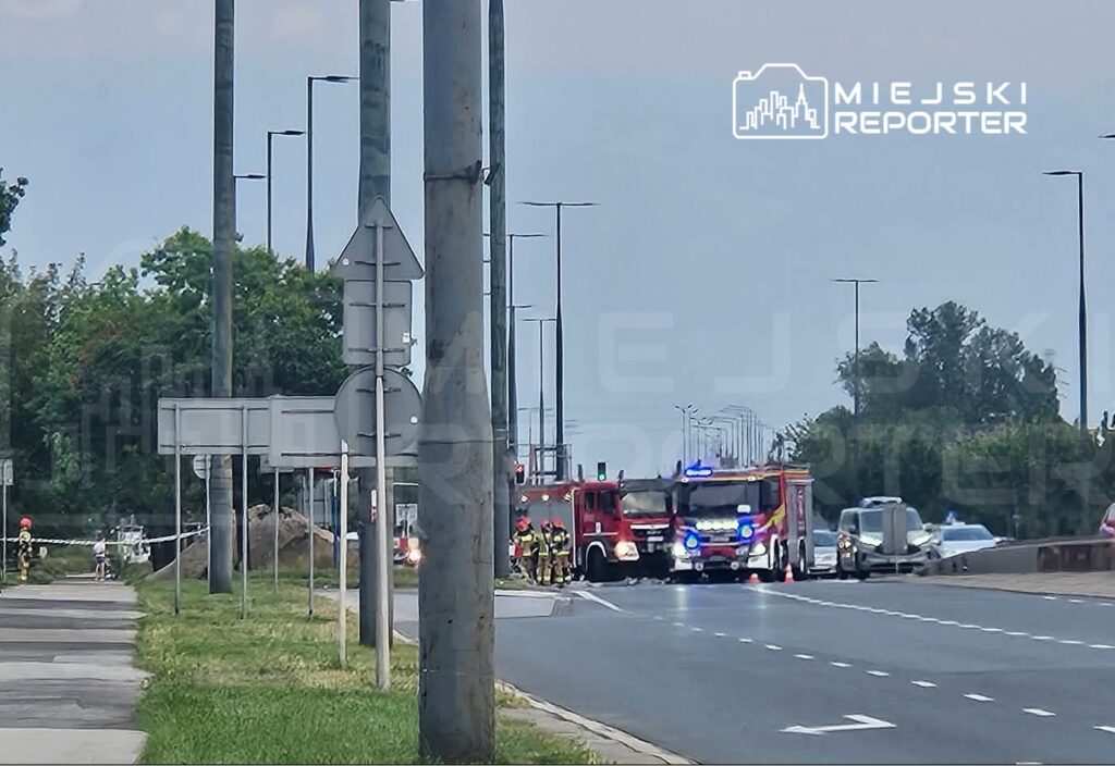 Na drodze stoi wóz strażacki z włączonymi sygnałami, a obok grupka strażaków zabezpiecza miejsce zdarzenia.