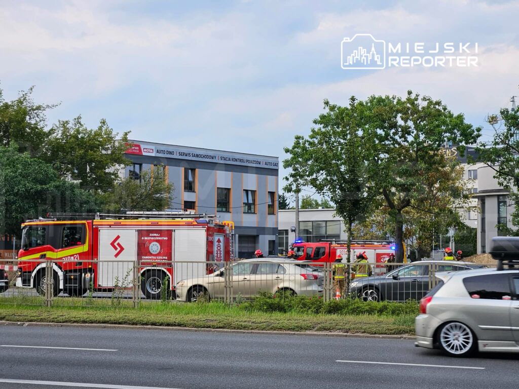 W pobliżu nowoczesnego biurowca z przeszkloną fasadą, samochody straży pożarnej interweniują na miejscu zdarzenia.