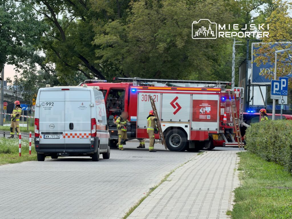 Strażacy w mundurach pracują przy wozie strażackim, obok karetki pogotowia, na miejskiej ulicy.