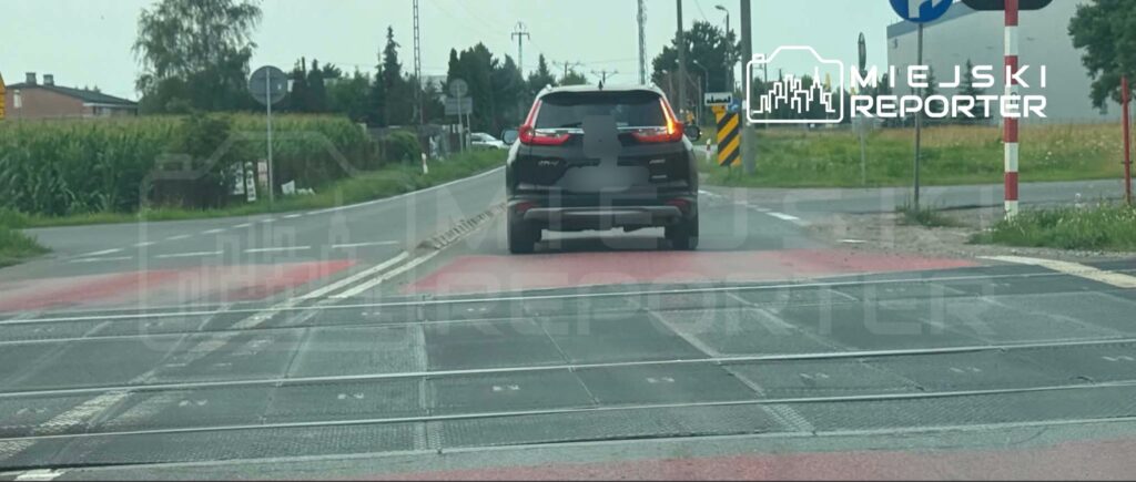 Czarny SUV przejeżdża przez skrzyżowanie z torami kolejowymi w otoczeniu pól i znaków drogowych.