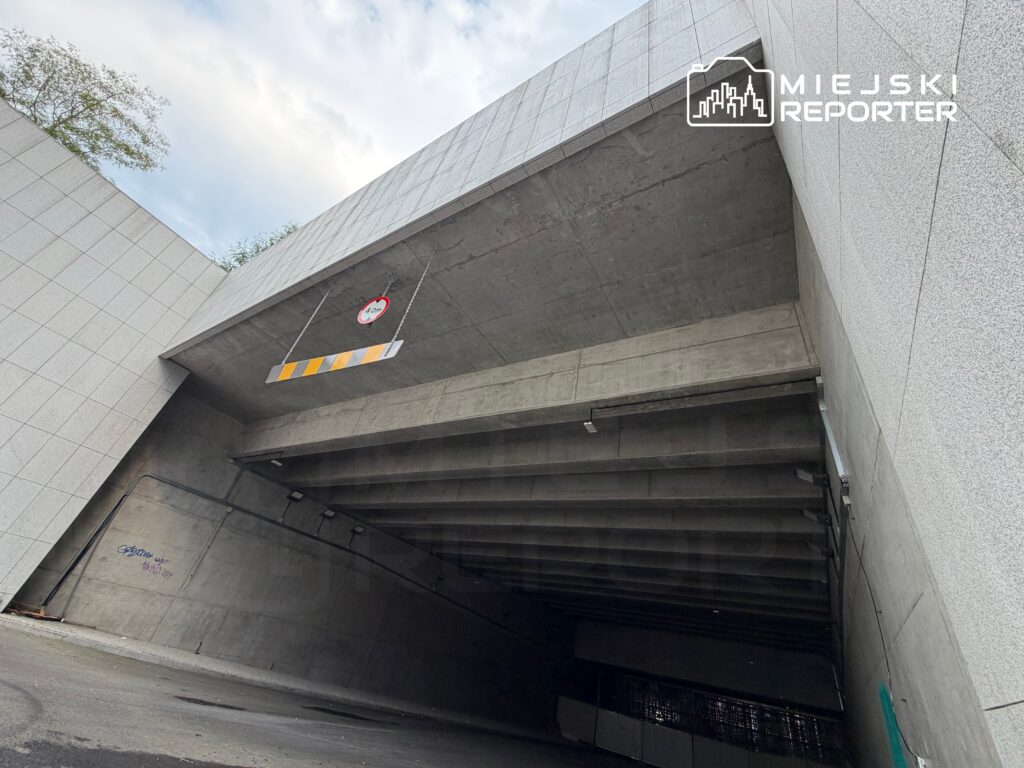 Wysoki, betonowy wjazd do parkingu podziemnego z oznaczeniem wysokości 2,10 m oraz graffiti na ścianie.
