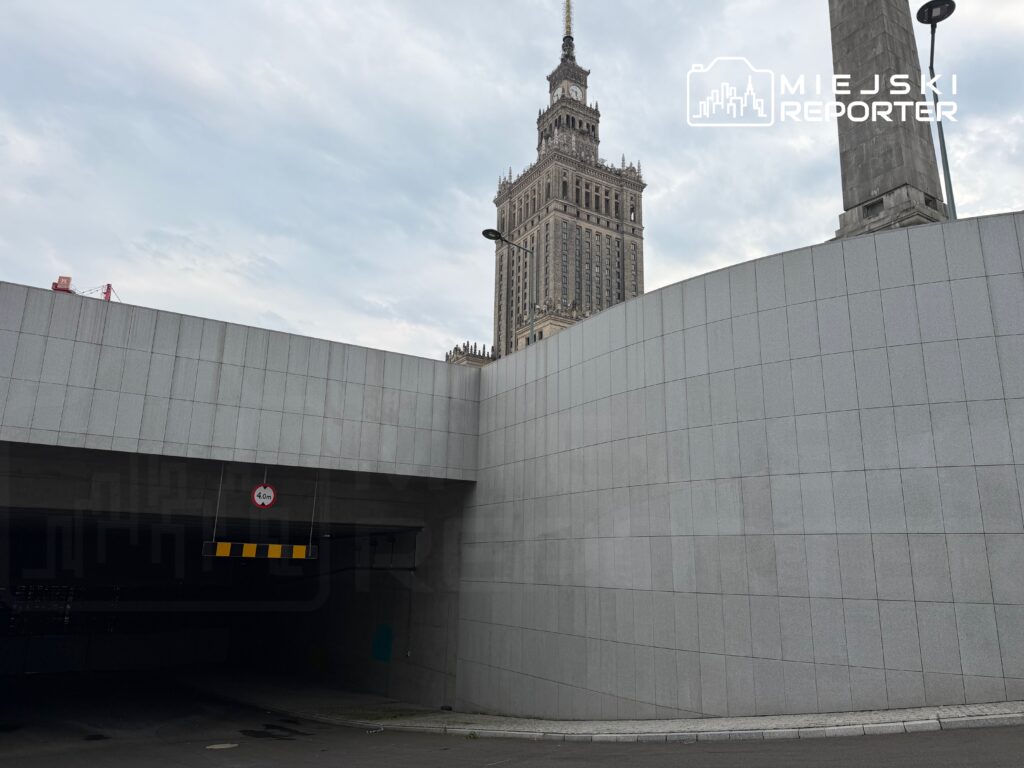 Wysoki budynek z zegarem w tle, nad wjazdem do podziemnego parkingu widoczny znak ograniczenia wysokości 4 m.