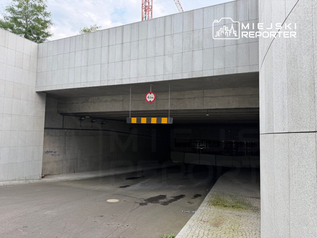 Wjazd do podziemnego parkingu z oznaczeniem wysokości 4 m, otoczony nowoczesnymi, szarymi ścianami.