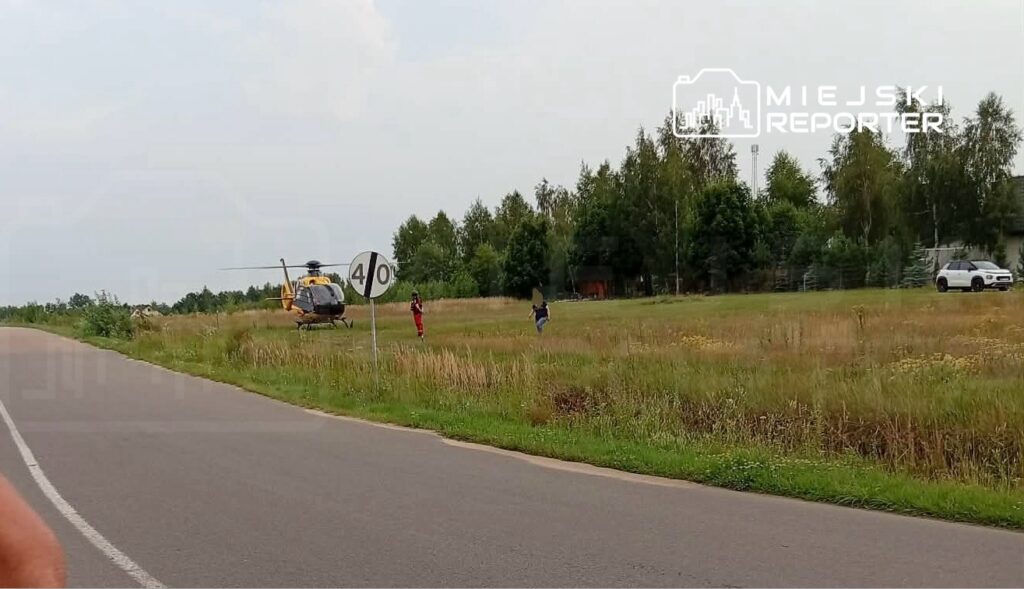 Helikopter lądował na łące obok drogi, a ratownicy rozmawiają z osobą stojącą w pobliżu znaku drogowym.