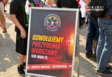 Chłopiec trzymający flagę stoi obok tablicy z hasłem "ODWOŁUJEMY PREZYDENTA WARSZAWY" na tle zgromadzenia ludzi.