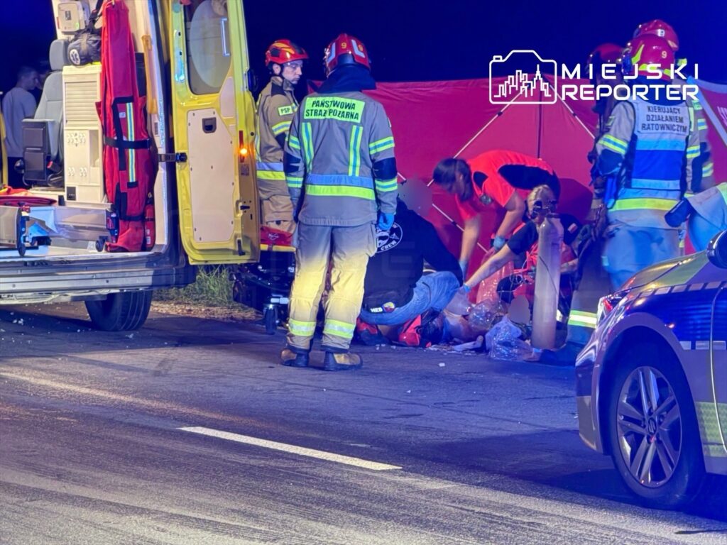 Grupa ratowników medycznych i strażaków udziela pomocy poszkodowanemu na drodze, otoczona sprzętem ratunkowym.