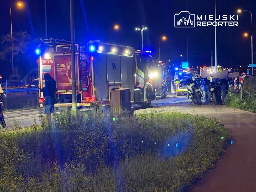 Grupa ratowników przy wozie strażackim na drodze, otoczeni pojazdami służb ratunkowych w nocy.