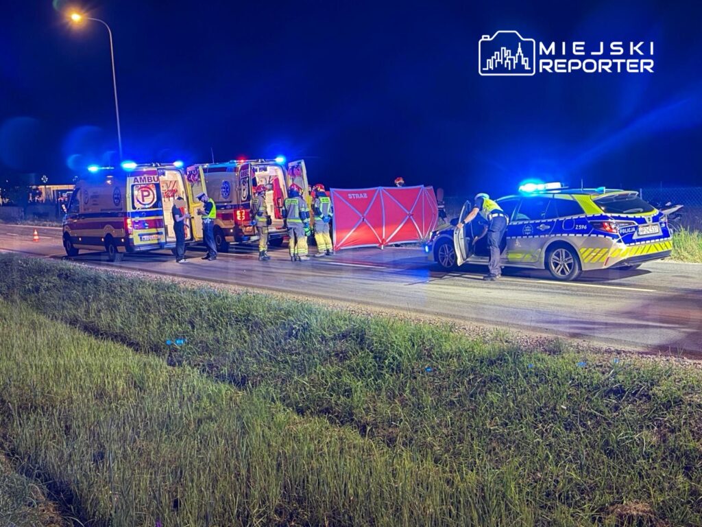 Na drodze nocą, zespół ratunkowy i policja pracują przy wypadku, używając ambulansów i sprzętu zabezpieczającego.