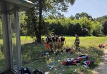 Grupa strażaków w mundurach z narzędziami w rękach przechodzi przez trawiaste pole, obok porozrzucane torby i sprzęt ratunkowy.