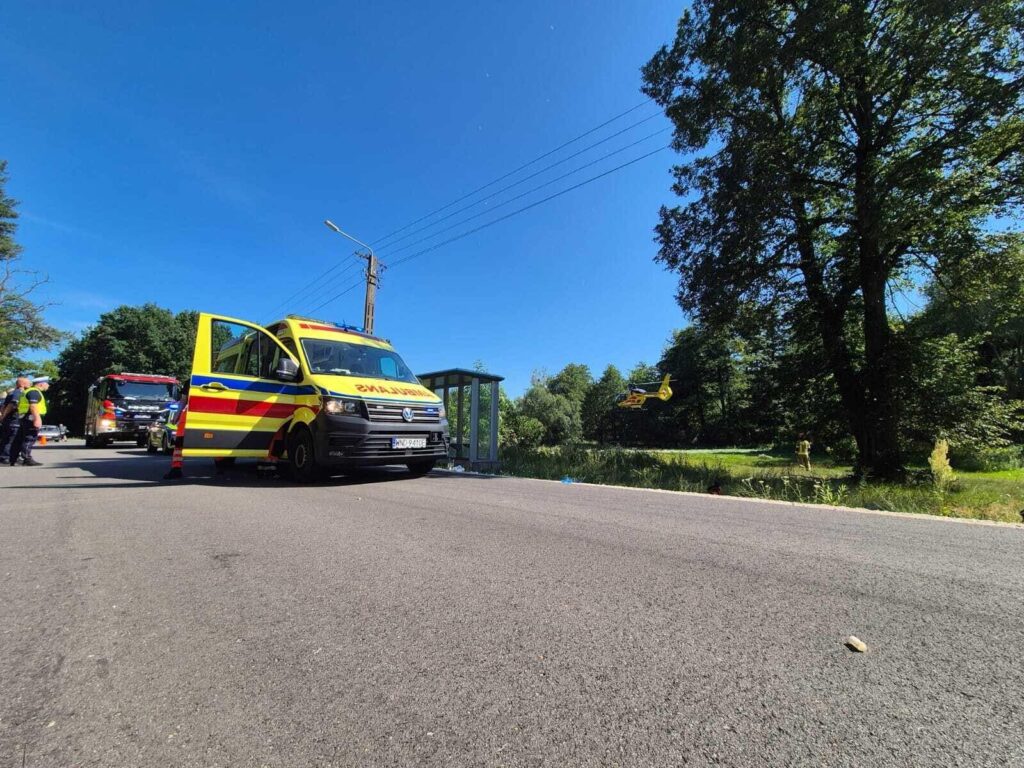 Zespół ratunkowy przy ambulansie na drodze, obok lądowa śmigłowca ratunkowego, w otoczeniu zieleni.