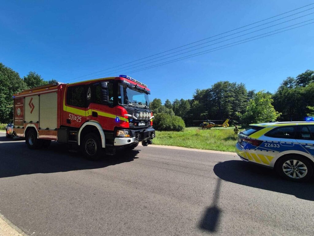 Wozidło strażackie zatrzymane na drodze, obok radiowozu policji, w tle lądowanie śmigłowca ratunkowego w lesie.