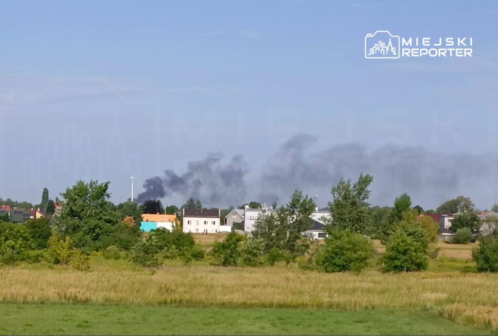 Czarny dym unoszący się nad zabudowaniami mieszkalnymi w otoczeniu zieleni i pól.