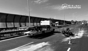 Na drodze leży przewrócony motocykl otoczony pachołkami, obok stoi laweta z podniesioną platformą.