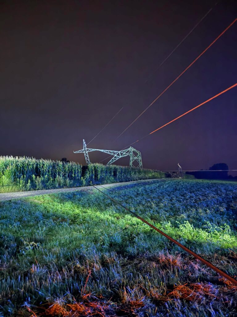 Wieś nocą z widocznym słupem wysokiego napięcia i liniami energetycznymi nad polem uprawnym.