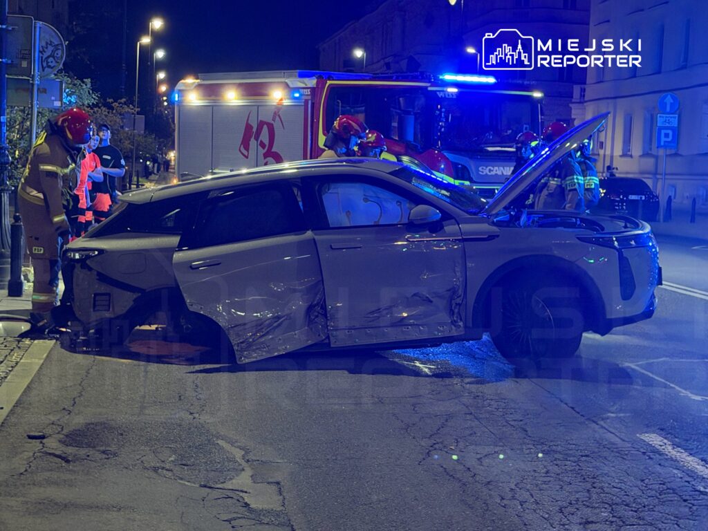 W nocy ratownicy medyczni i strażacy udzielają pomocy przy uszkodzonym pojeździe na ulicy.