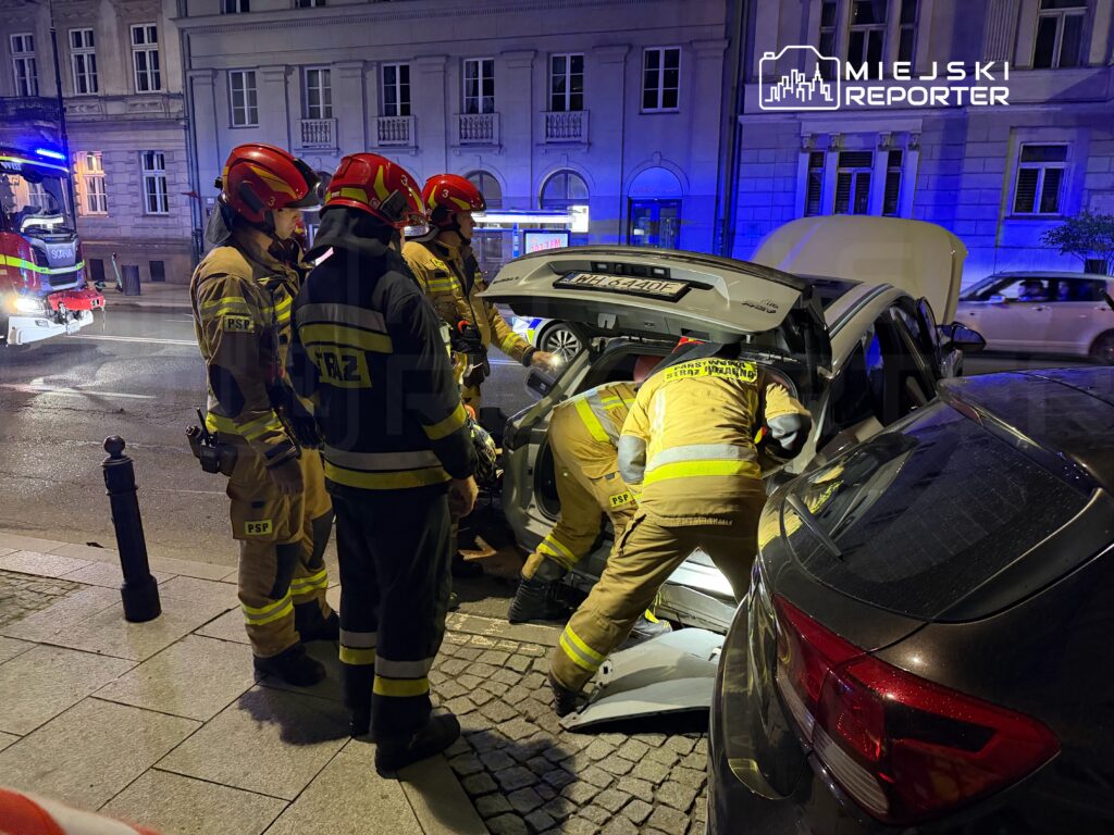 Grupa strażaków w żółtych mundurach pracuje przy otwartym bagażniku samochodu na nocnej ulicy.