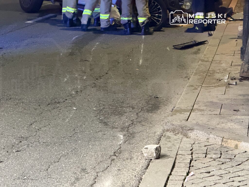Grupa strażaków udziela pomocy poszkodowanym na jezdni w miejskim otoczeniu, obok uszkodzonego pojazdu.