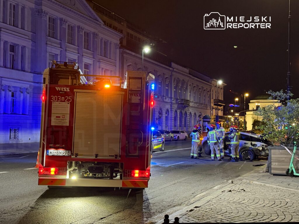 Strażacy w specjalistycznym sprzęcie prowadzą działania ratunkowe przy uszkodzonym samochodzie na ulicy nocą.