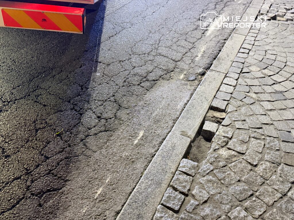 Zniszczona nawierzchnia drogi z widocznymi pęknięciami i uszkodzeniami, obok kostka brukowa.