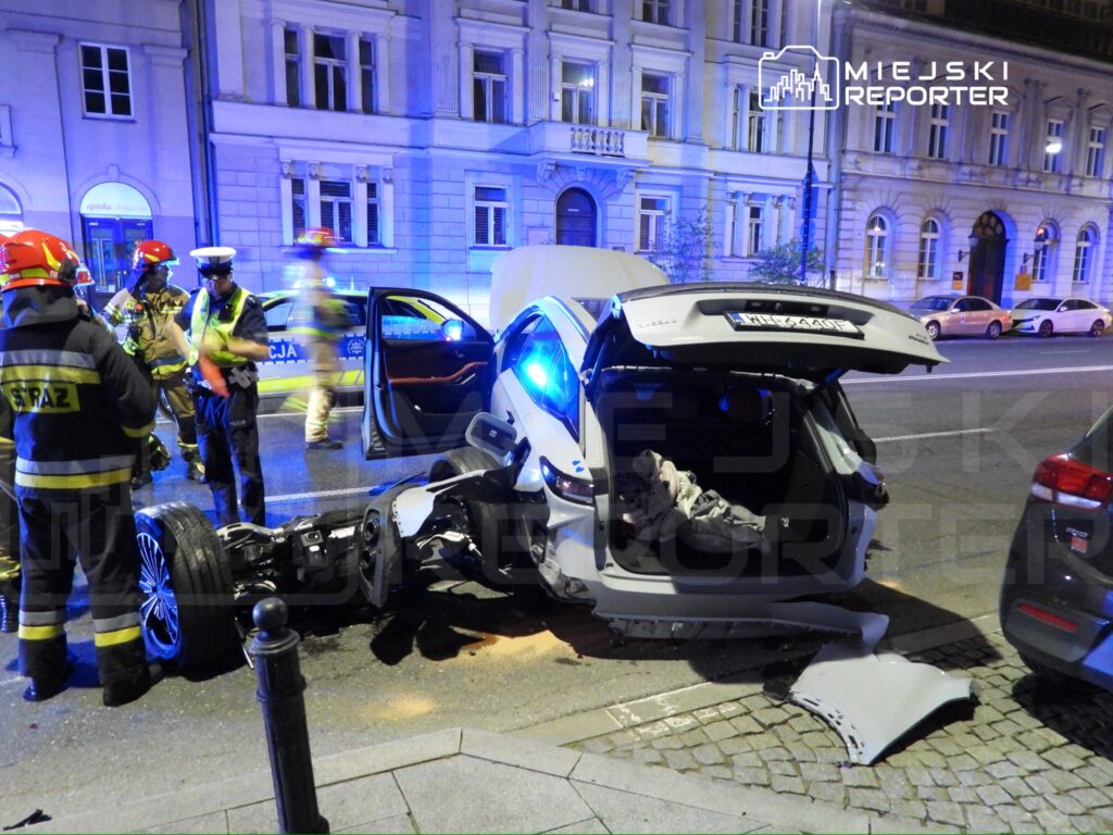 W nocy, na ulicy, strażacy i policjanci pracują przy rozbitej, uszkodzonej na wylot, białej limuzynie.