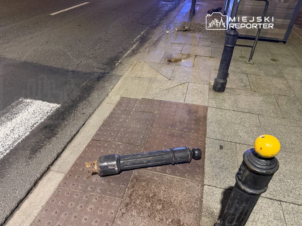 Złamany słupek oświetleniowy leży na chodniku, obok prostokątnej płyty chodnikowej i krawężnika.