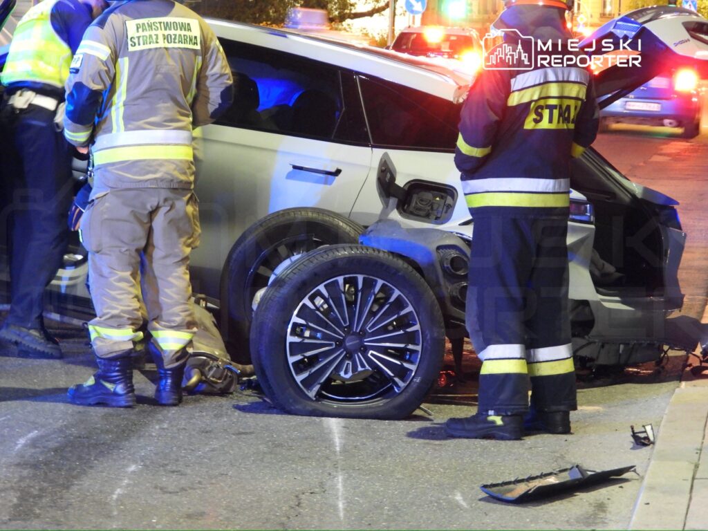 Strażacy w mundurach interweniują przy uszkodzonym samochodzie na ulicy, obok leży odłamany fragment pojazdu.