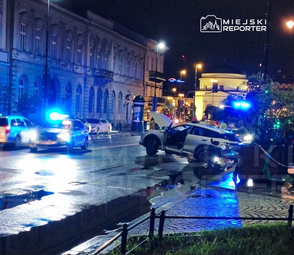 W nocy na ulicy widać uszkodzony samochód osobowy z otwartymi drzwiami, otoczony przez policyjne radiowozy z włączonymi sygnałami świetlnymi.