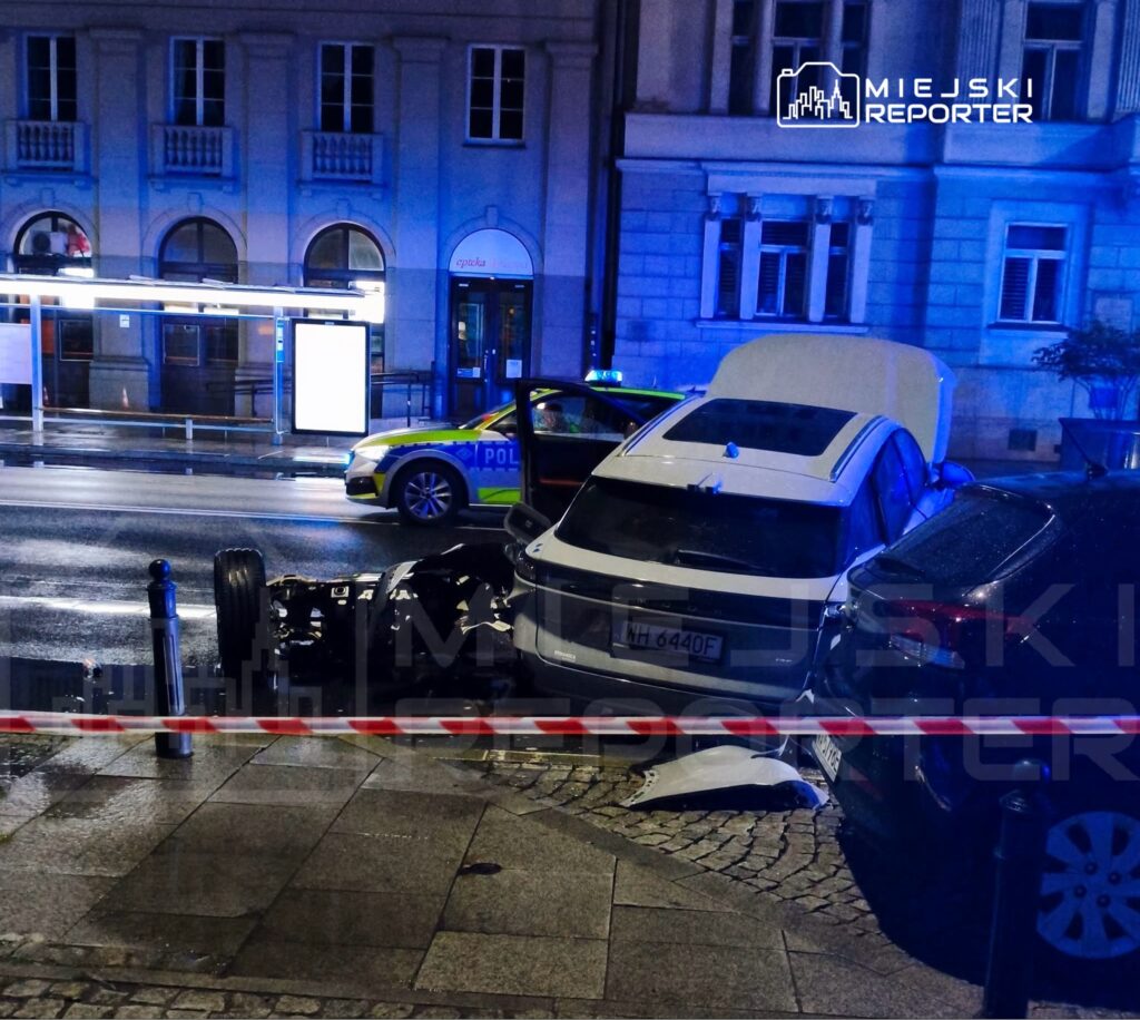 Uszkodzony samochód osobowy z oderwanym kołem stoi na chodniku, obok radiowóz policyjny z włączonymi światłami.