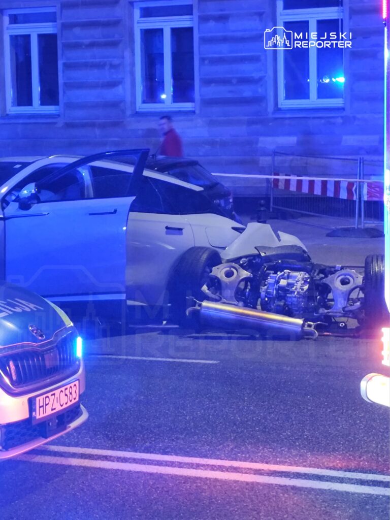 Uszkodzony samochód osobowy z oderwaną tylną częścią stoi na ulicy, obok radiowóz policyjny z włączonymi sygnałami świetlnymi.