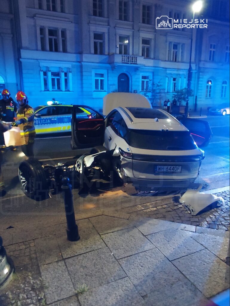 Samochód osobowy z uszkodzoną tylną częścią stoi na ulicy, obok interweniują strażacy oraz radiowóz policyjny.