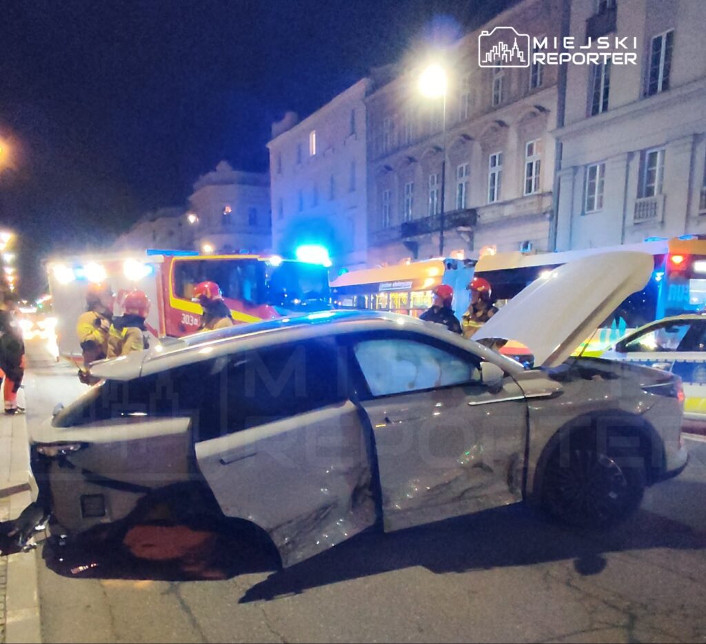 Na ulicy, w nocy, strażacy z włączonymi sygnalizacjami pomagają przy uszkodzonym samochodzie osobowym.