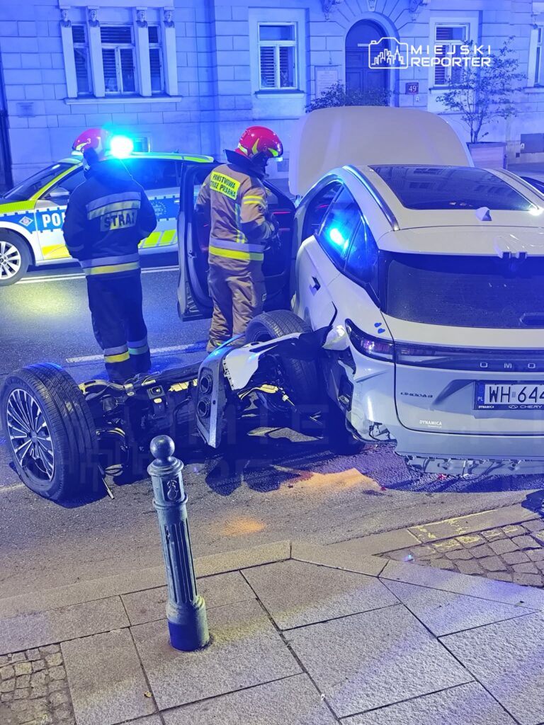 Dwaj strażacy w kaskach badają uszkodzony samochód osobowy na ulicy, obok policyjny radiowóz z włączonymi sygnałami.