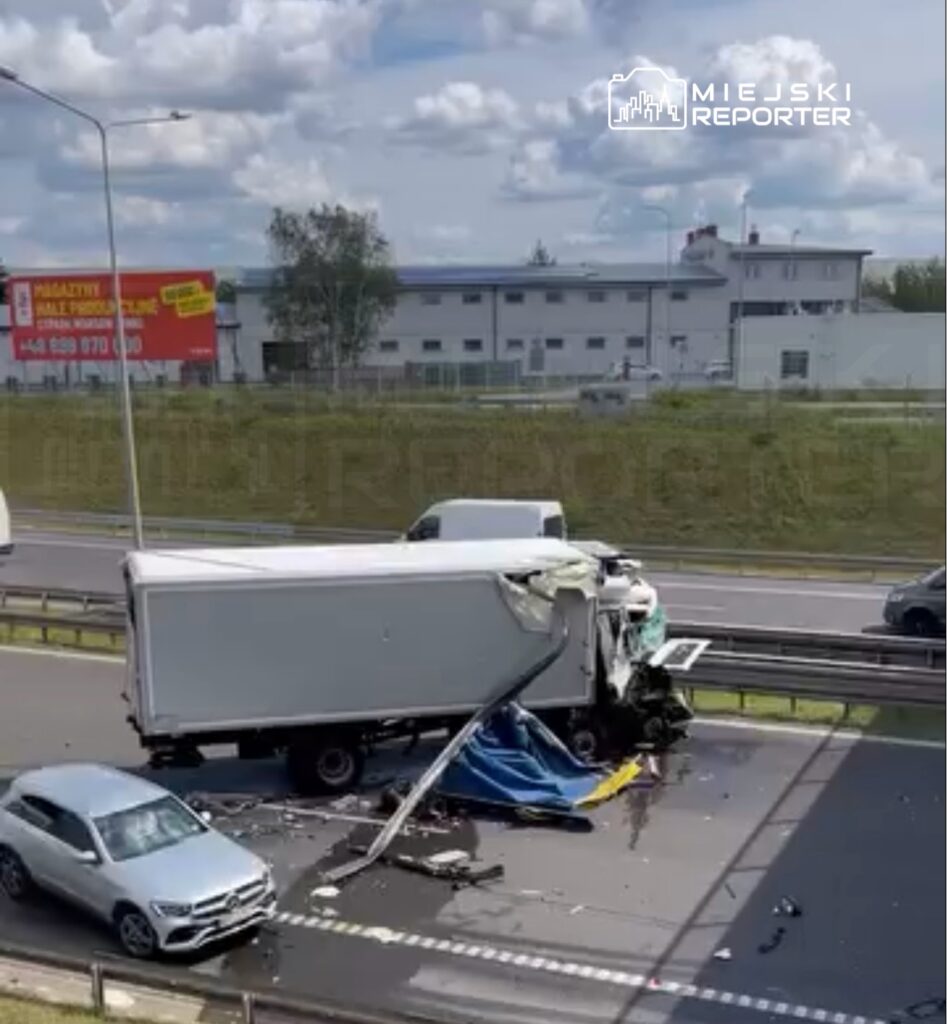 Zniszczony samochód ciężarowy z uszkodzoną kabiną oraz rozrzuconymi elementami na drodze, obok osobowy samochód.
