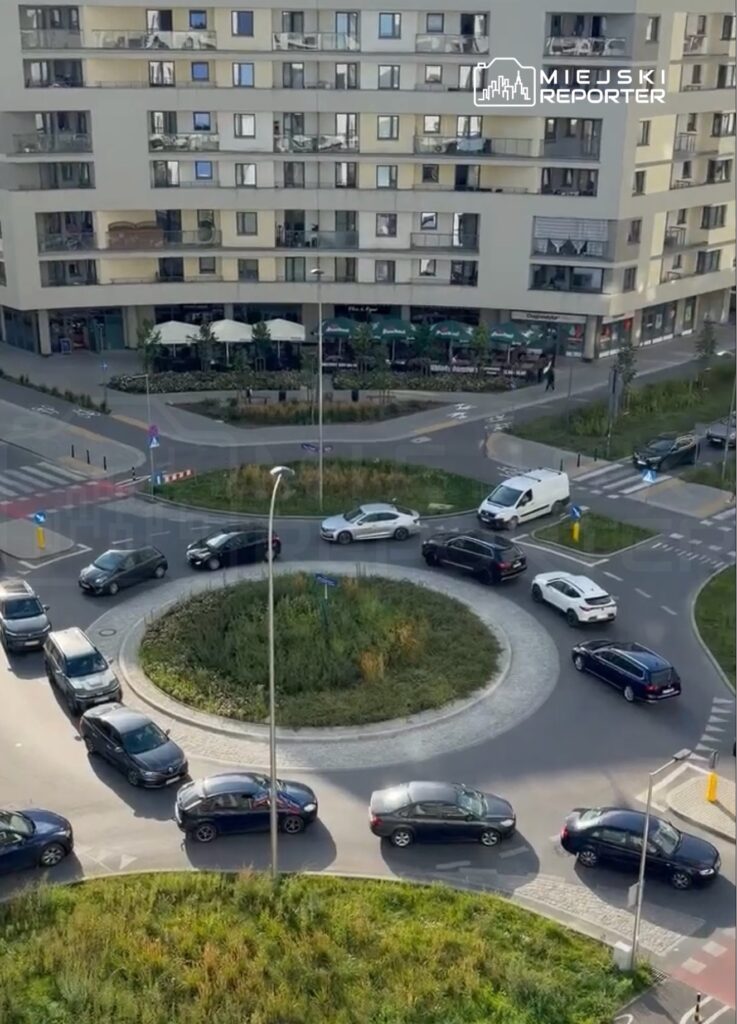 Ruch samochodów na rondzie w nowoczesnej okolicy, z widocznymi budynkami i restauracjami w tle.