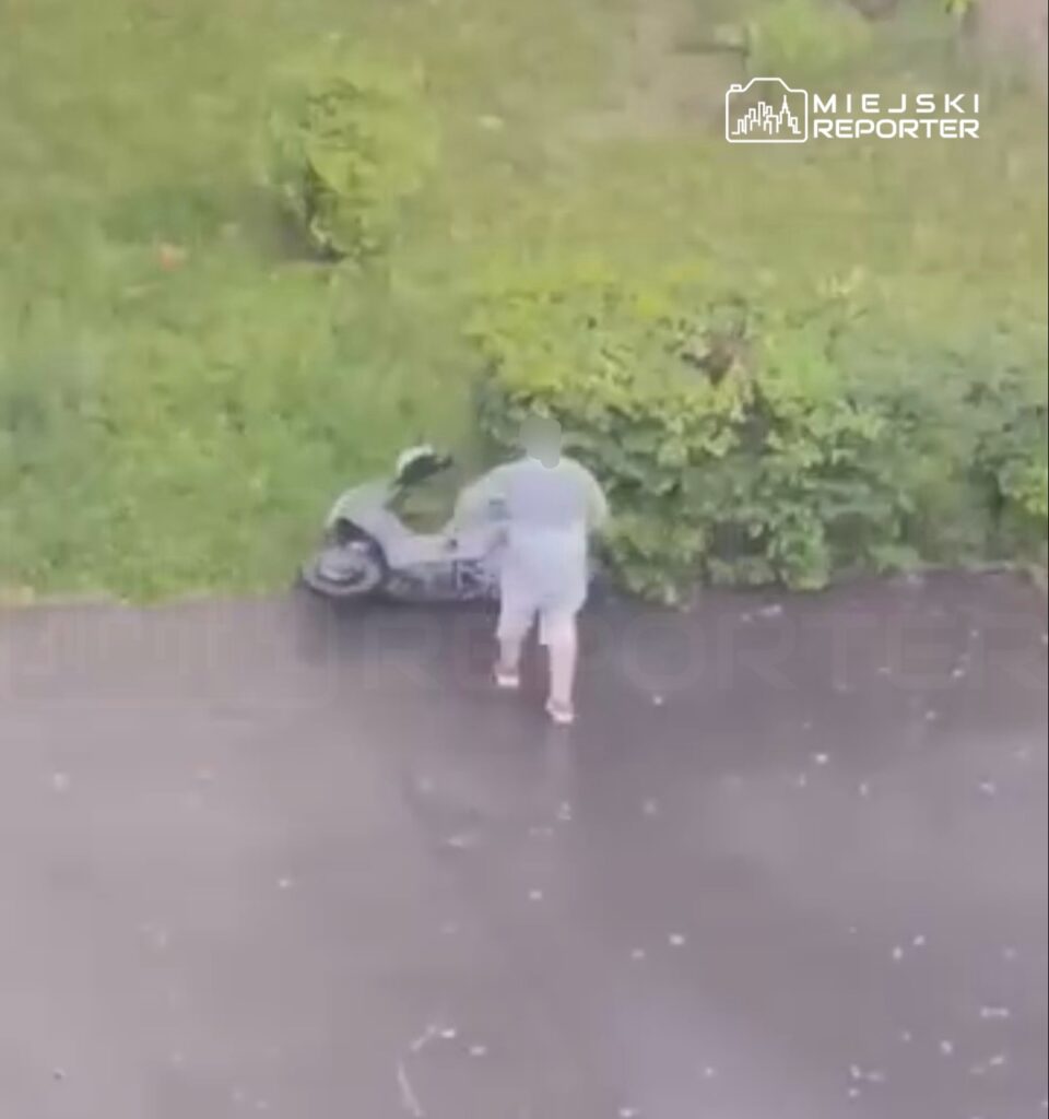 Mężczyzna w szarym stroju sprawdza skuter zaparkowany obok krzewów na asfaltowej nawierzchni.