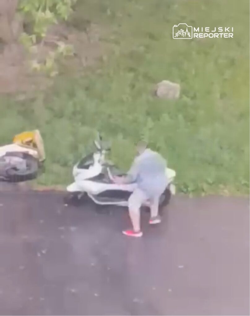 Mężczyzna w szarej odzieży przymierza się do uruchomienia skuteru zaparkowanego na trawniku obok asfaltowej drogi.