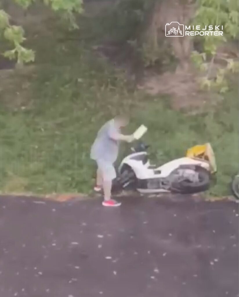 Mężczyzna w szarej koszulce trzyma kartkę i pochyla się nad białym skuterem zaparkowanym na trawie.