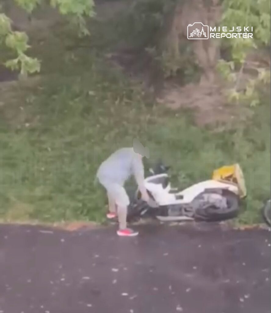 Mężczyzna w szarej odzieży podnosi przewrócony skuter na trawie obok drogi.