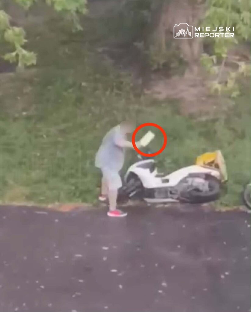 Mężczyzna w szarej koszulce podnosi przedmiot z trawnika obok uszkodzonego skutera.