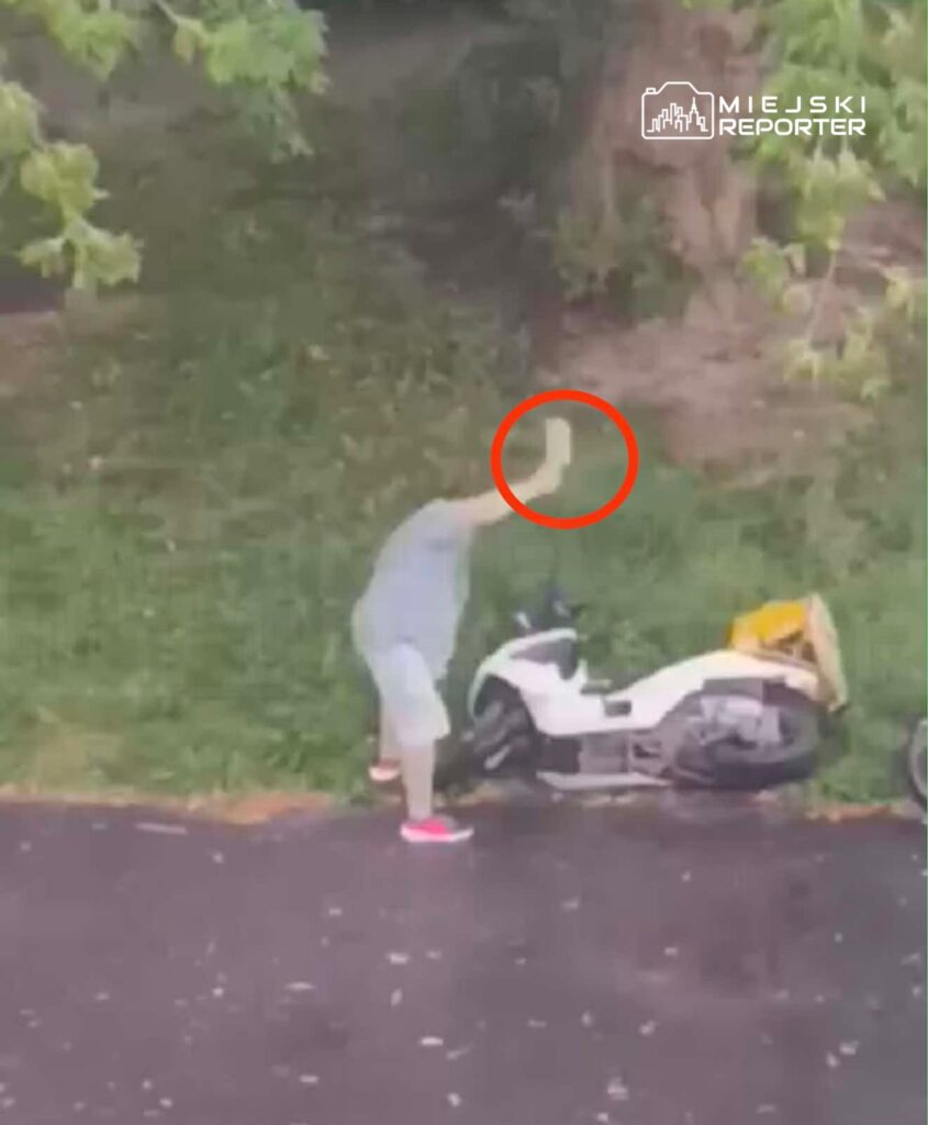 Mężczyzna w szarej odzieży podnosi rękę w kierunku motocykla zaparkowanego na trawie obok drogi.