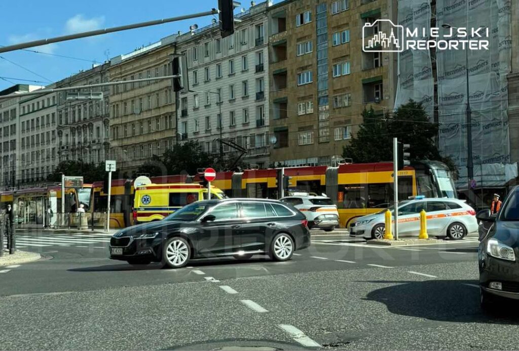 Czarny samochód osobowy skręca w lewo na ruchliwym skrzyżowaniu, obok tramwajów i karetek pogotowia.