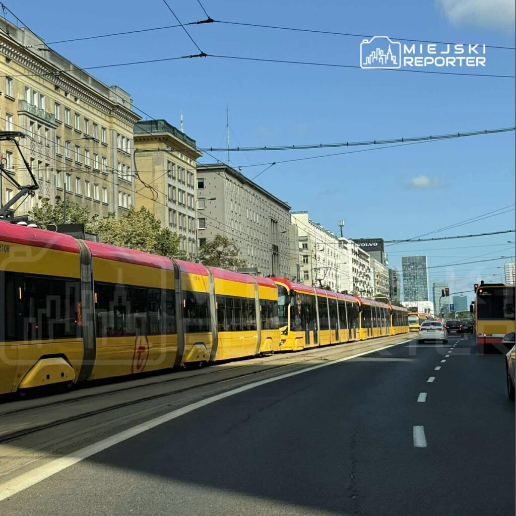 Tramwaj z różowym dachem porusza się po torach wzdłuż ruchliwej ulicy w mieście, otoczony budynkami.