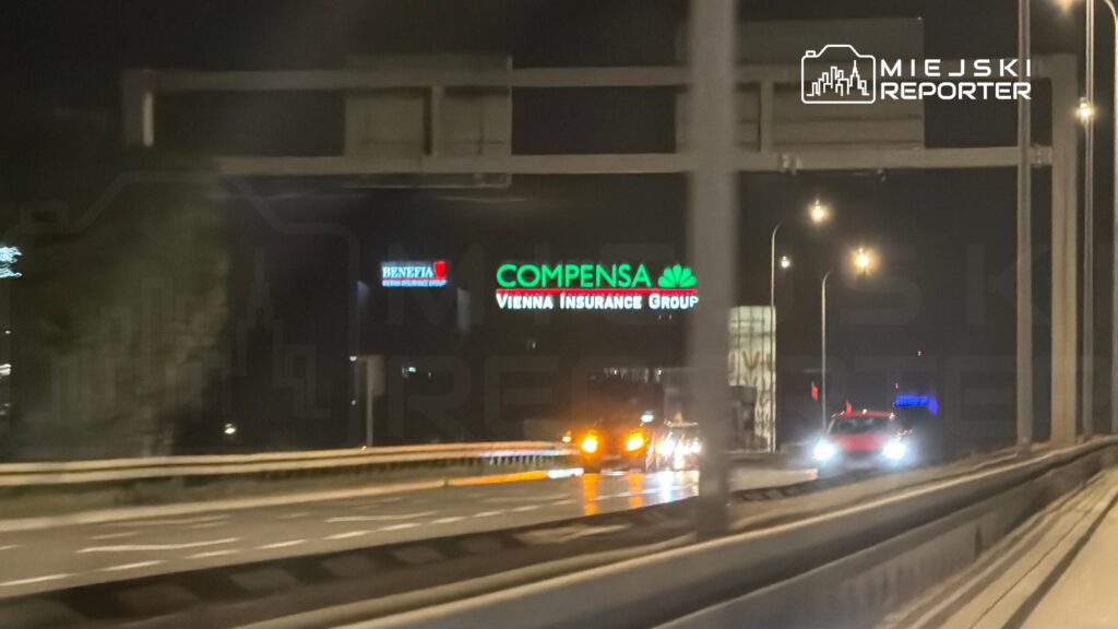 Na drodze nocą poruszają się samochody, obok widoczne podświetlone reklamy firm ubezpieczeniowych.