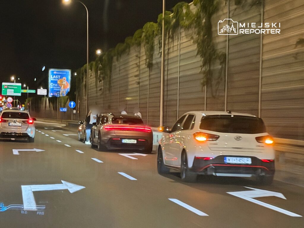 Trzy samochody poruszają się nocą po drodze, jeden z nich zatrzymuje się obok innego, na tle billboardów.