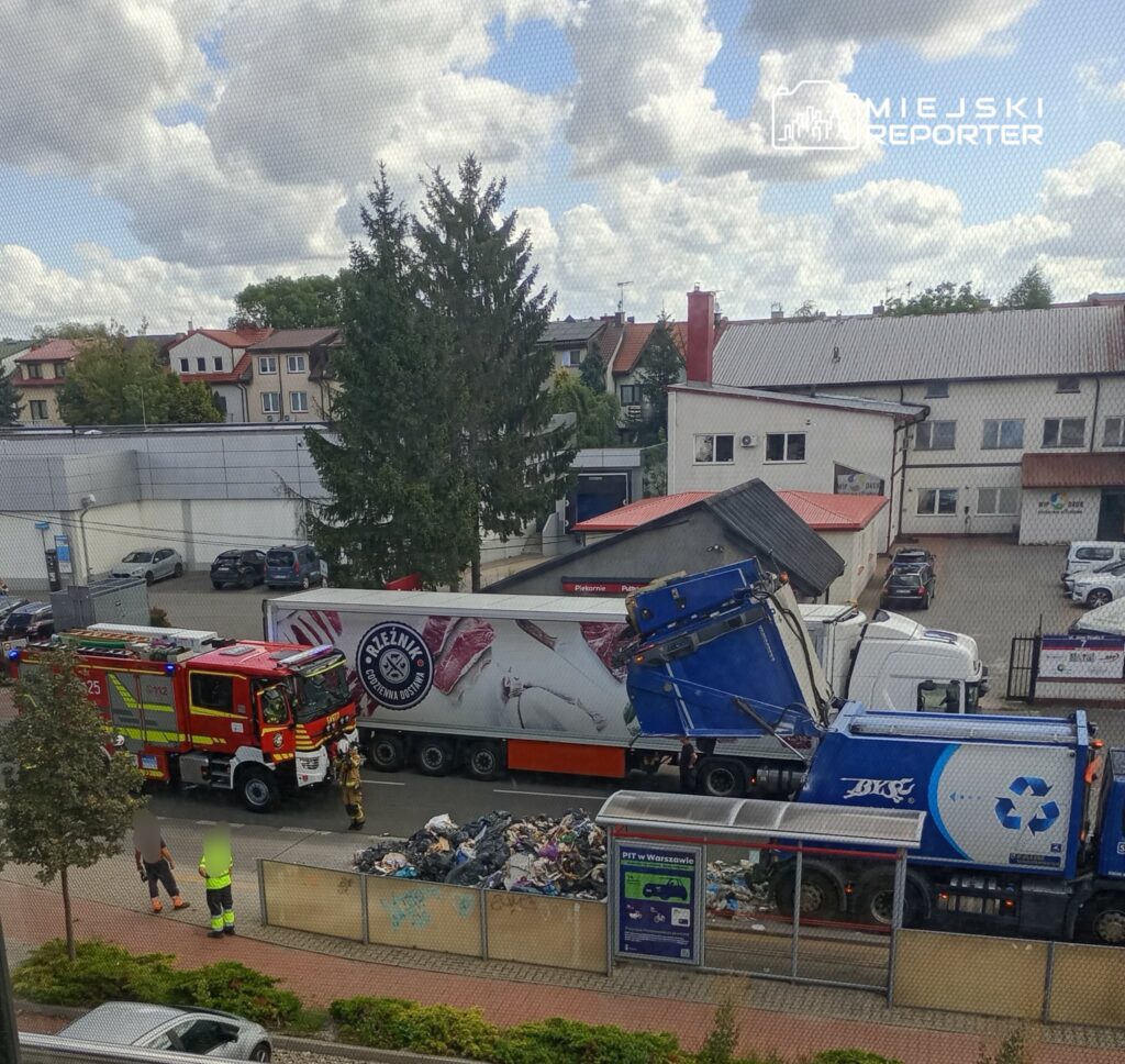 Wóz strażacki oraz śmieciarka stoją na ulicy, obok wypełnionego odpadkami kontenera, w tle budynki mieszkalne.