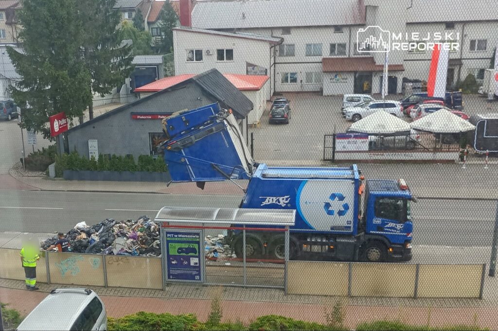 Śmieciarka opróżniająca kontener z odpadami na ulicy, obok budynków i przystanku autobusowego.