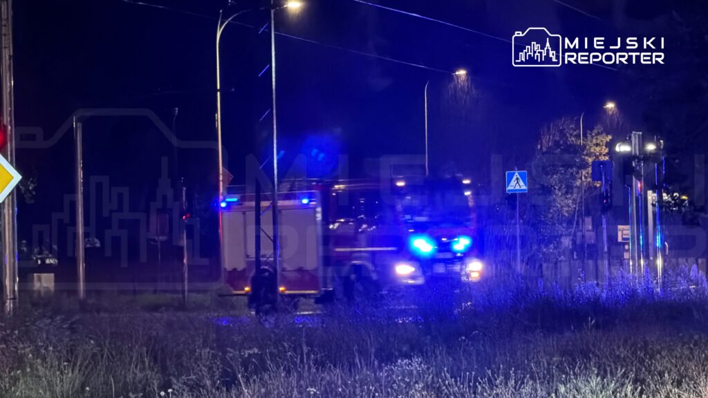 Karetka pogotowia z włączonymi sygnałami świetlnymi przejeżdża przez ciemną ulicę, obok znaku drogowym.