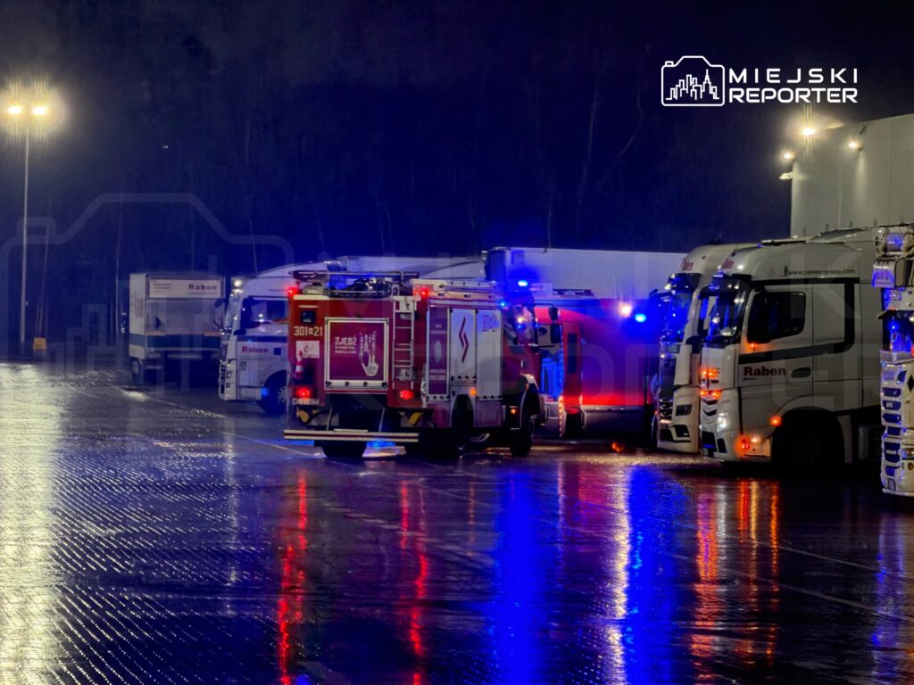W nocy na parkingu ciężarówek, widać pojazd strażacki z włączonymi sygnalizacjami świetlnymi obok zaparkowanych tirów.