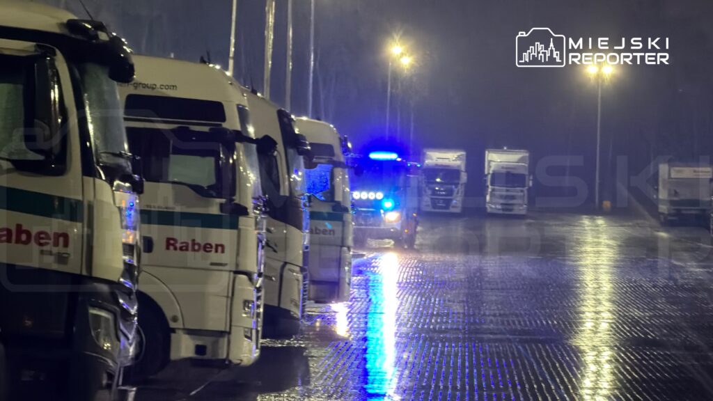Tiry zaparkowane na oświetlonym parkingu w deszczową noc, w tle widoczne światła policyjnego pojazdu.